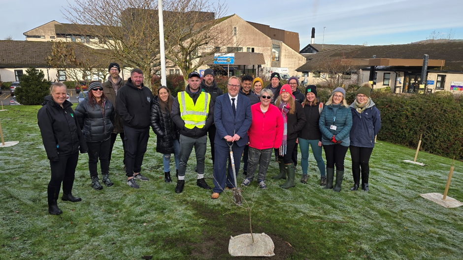 A group of people standing on grass outside Westmorland General Hospital in Kendal with spades and surrounded by trees